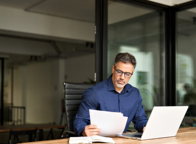 Busy mature professional business man using laptop reading document at work.