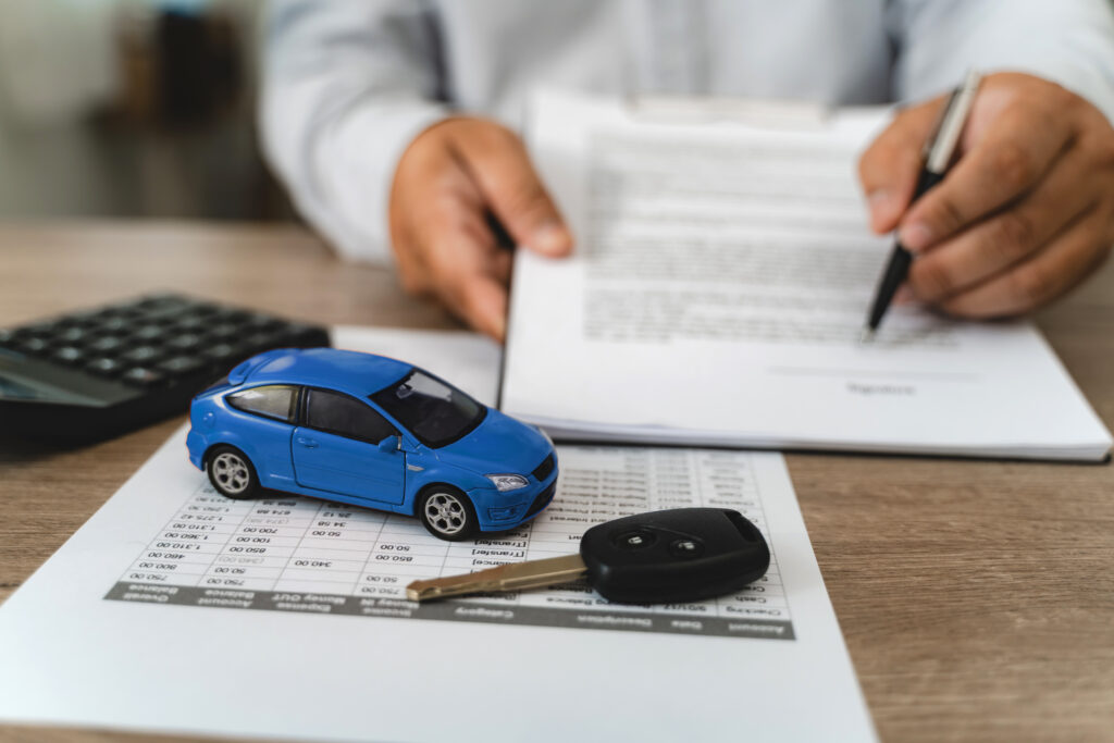 Hands reviewing car loan finance document contract with payment schedule table, blue toy car model and key on paperwork, calculator on desk, representing auto finance agreement analysis, vehicle lease payment plan review