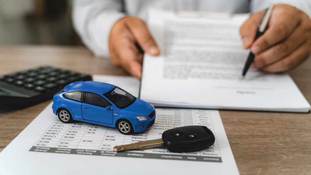 Hands reviewing car loan finance document contract with payment schedule table, blue toy car model and key on paperwork, calculator on desk, representing auto finance agreement analysis, vehicle lease payment plan review