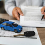 Hands reviewing car loan finance document contract with payment schedule table, blue toy car model and key on paperwork, calculator on desk, representing auto finance agreement analysis, vehicle lease payment plan review