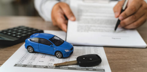 Hands reviewing car loan finance document contract with payment schedule table, blue toy car model and key on paperwork, calculator on desk, representing auto finance agreement analysis, vehicle lease payment plan review