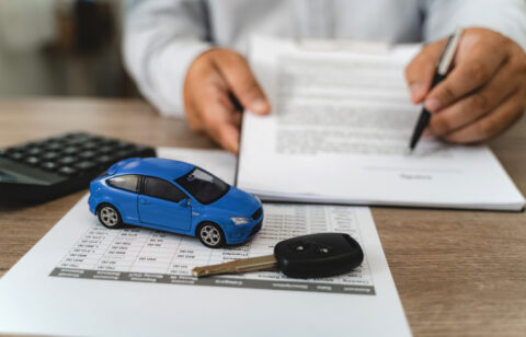 Hands reviewing car loan finance document contract with payment schedule table, blue toy car model and key on paperwork, calculator on desk, representing auto finance agreement analysis, vehicle lease payment plan review