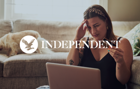 Woman sitting on a couch looking stressed while holding a credit card and using a laptop, with The Independent logo overlaid.