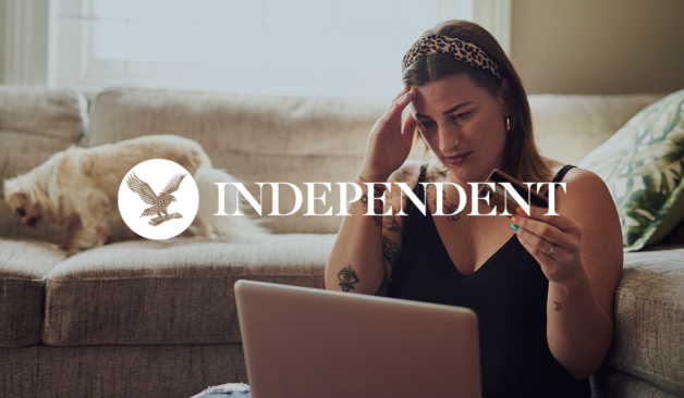 Woman sitting on a couch looking stressed while holding a credit card and using a laptop, with The Independent logo overlaid.