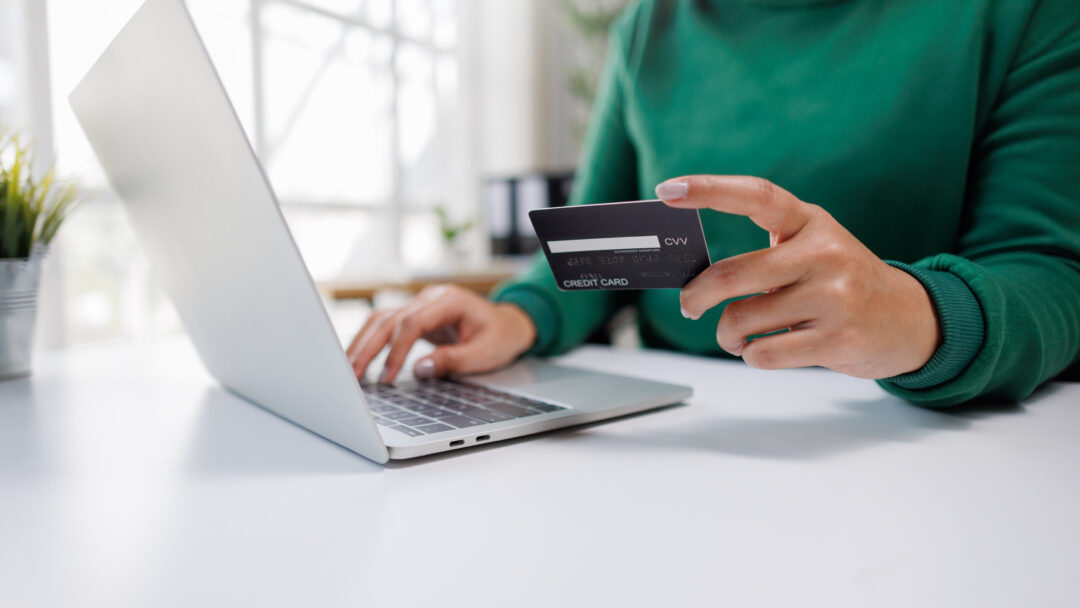 A person is engaged in online shopping, holding a credit card in one hand and typing on a laptop with the other, showcasing modern digital transaction habits.