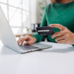 A person is engaged in online shopping, holding a credit card in one hand and typing on a laptop with the other, showcasing modern digital transaction habits.