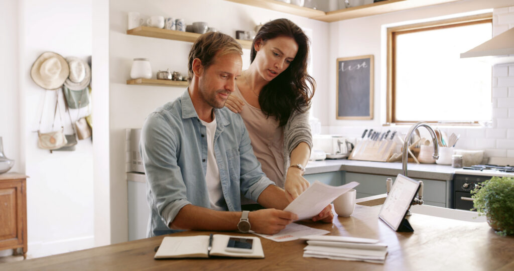 Man and woman reviewing financial paperwork together at a kitchen table with a tablet and notebook nearby.