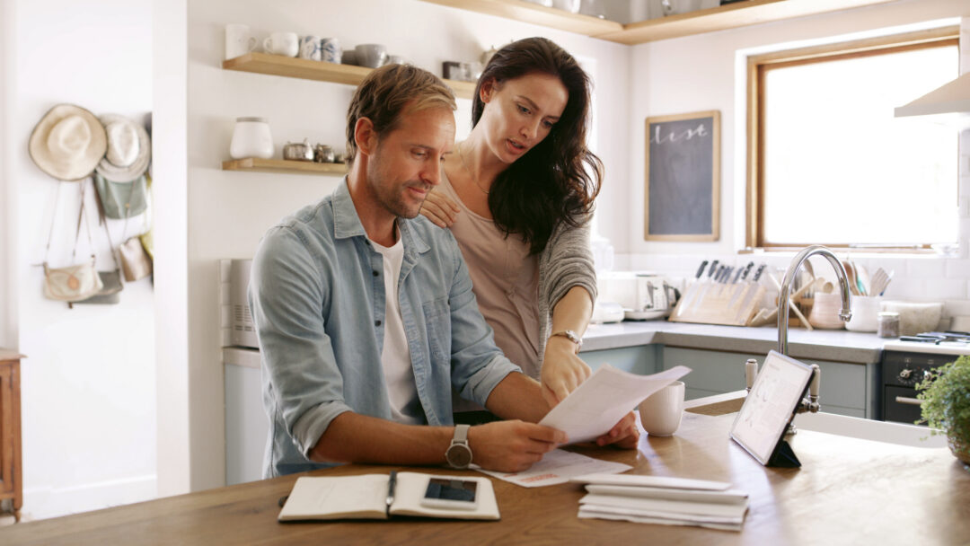 Man and woman reviewing financial paperwork together at a kitchen table with a tablet and notebook nearby.