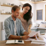 Man and woman reviewing financial paperwork together at a kitchen table with a tablet and notebook nearby.