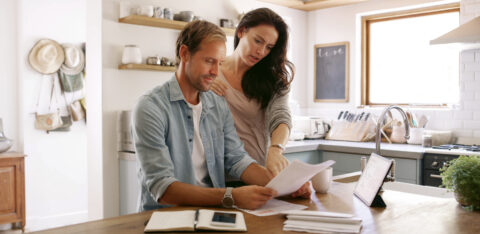 Man and woman reviewing financial paperwork together at a kitchen table with a tablet and notebook nearby.