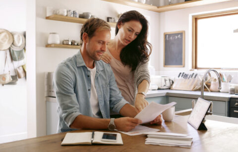 Man and woman reviewing financial paperwork together at a kitchen table with a tablet and notebook nearby.