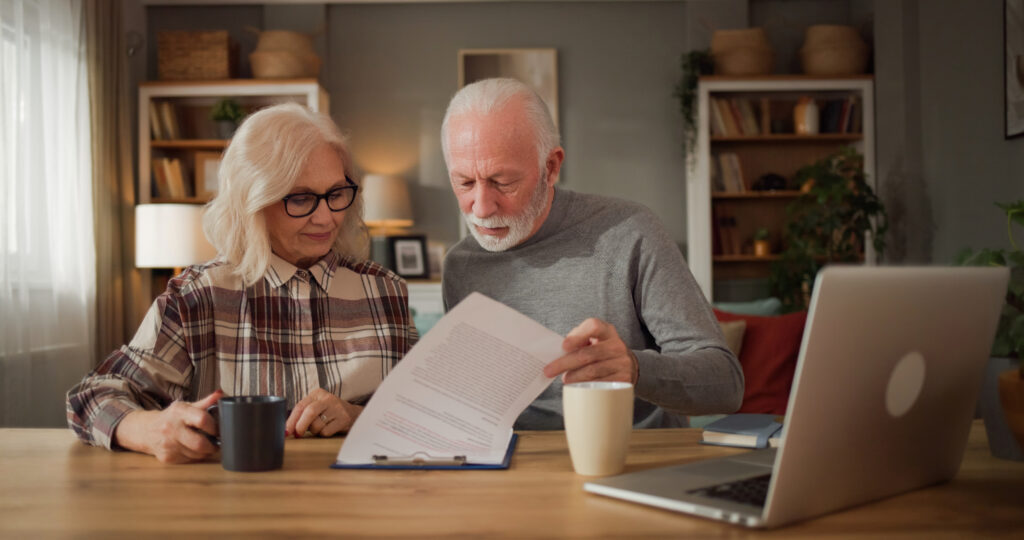 Older couple reading paperwork together at a wooden table with a laptop and coffee mugs.