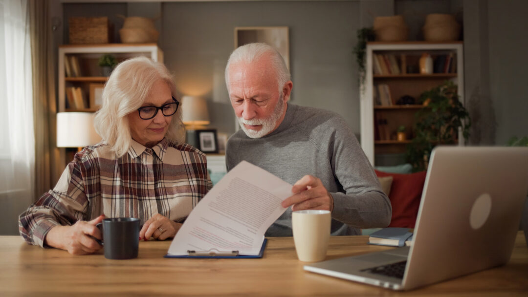 Older couple reading paperwork together at a wooden table with a laptop and coffee mugs.