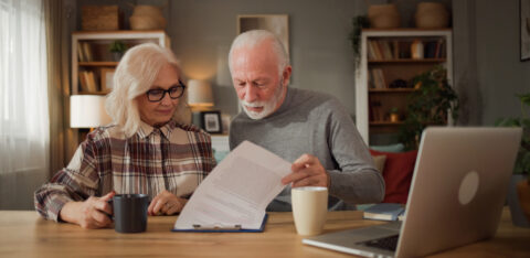 Older couple reading paperwork together at a wooden table with a laptop and coffee mugs.