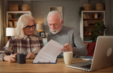 Older couple reading paperwork together at a wooden table with a laptop and coffee mugs.