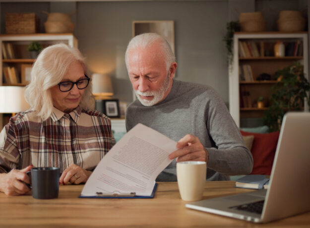 Older couple reading paperwork together at a wooden table with a laptop and coffee mugs.