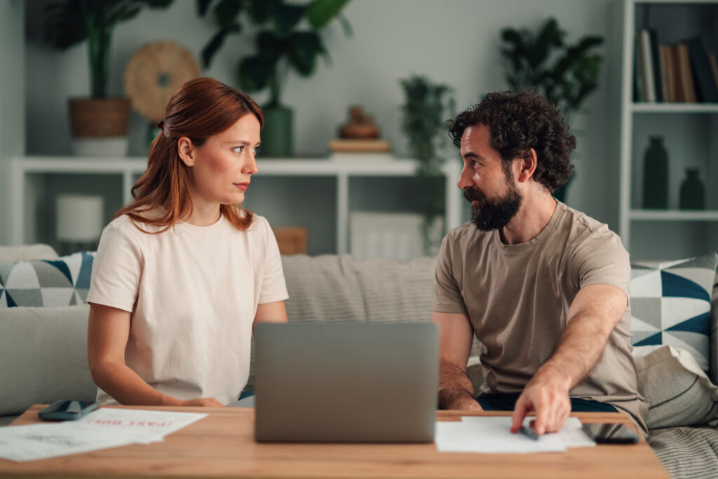 Couple sitting on a sofa with a laptop and papers, talking to each other.