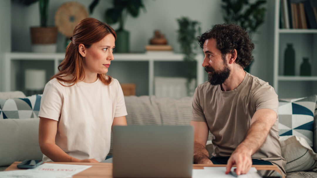 Couple sitting on a sofa with a laptop and papers, talking to each other.