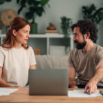 Couple sitting on a sofa with a laptop and papers, talking to each other.
