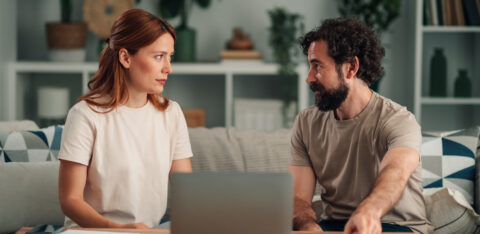 Couple sitting on a sofa with a laptop and papers, talking to each other.