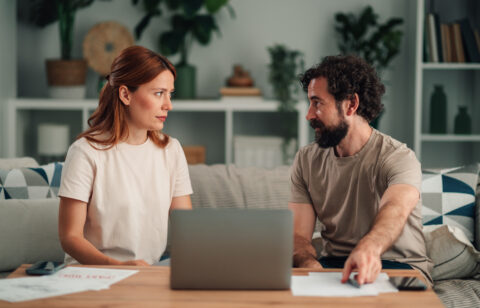 Couple sitting on a sofa with a laptop and papers, talking to each other.
