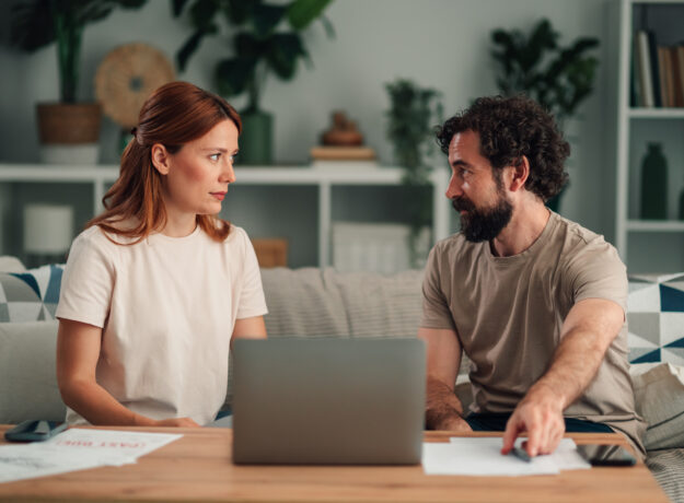 Couple sitting on a sofa with a laptop and papers, talking to each other.