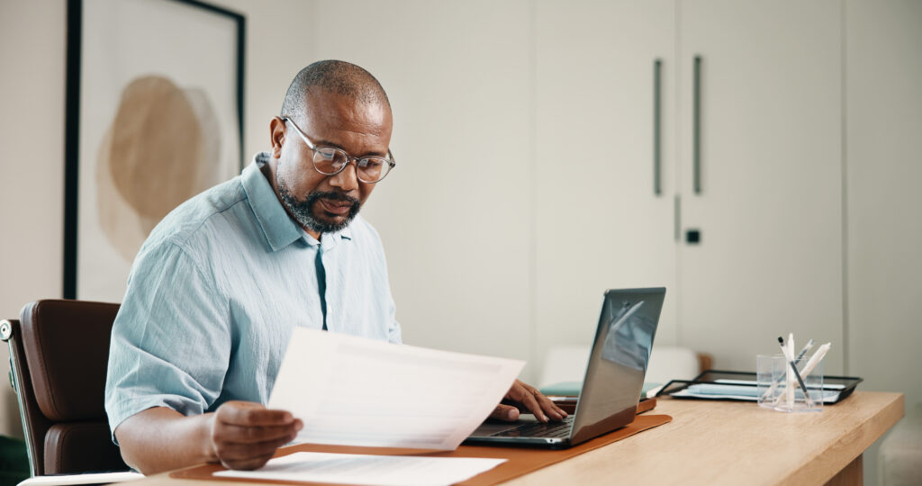 Man wearing glasses reviewing paperwork while working on a laptop at his desk.