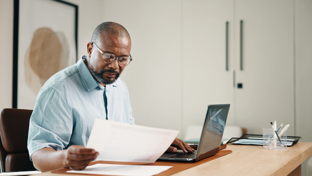 Man wearing glasses reviewing paperwork while working on a laptop at his desk.
