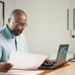 Man wearing glasses reviewing paperwork while working on a laptop at his desk.