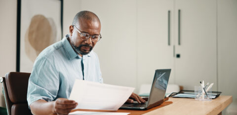 Man wearing glasses reviewing paperwork while working on a laptop at his desk.