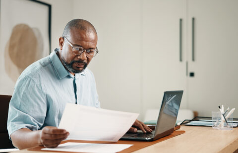 Man wearing glasses reviewing paperwork while working on a laptop at his desk.