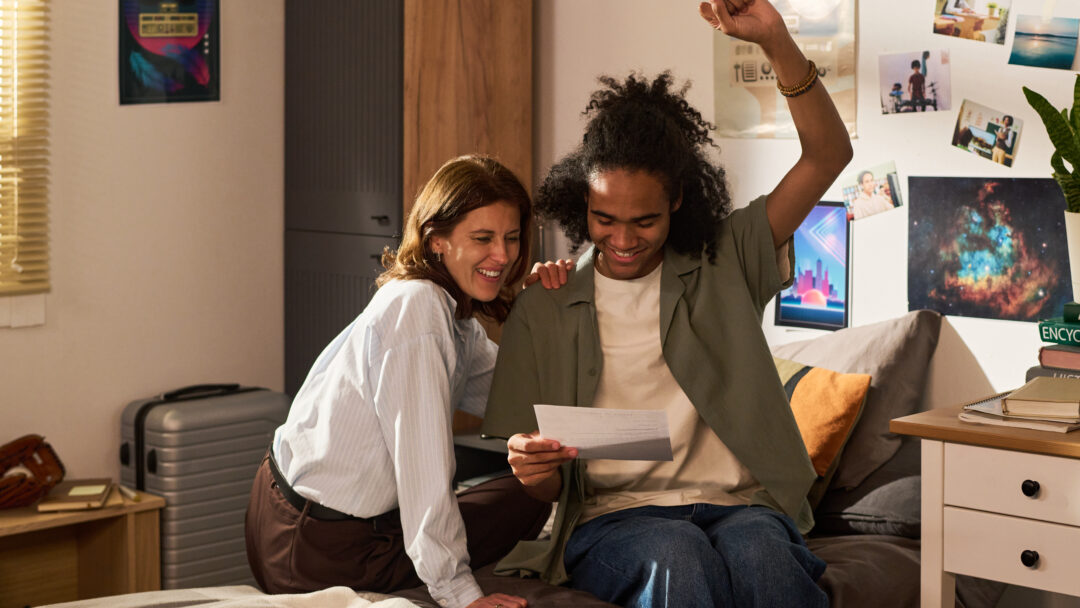 A college student raises his fist in excitement as his mother smiles beside him on a bed.