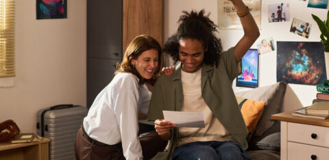 A college student raises his fist in excitement as his mother smiles beside him on a bed.