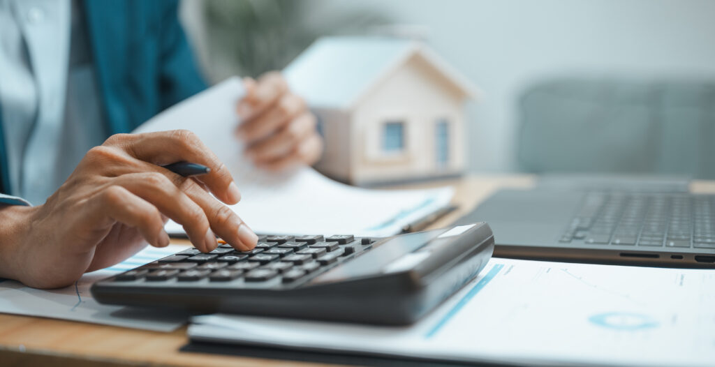 Person using a calculator while reviewing financial documents beside a laptop and model home.