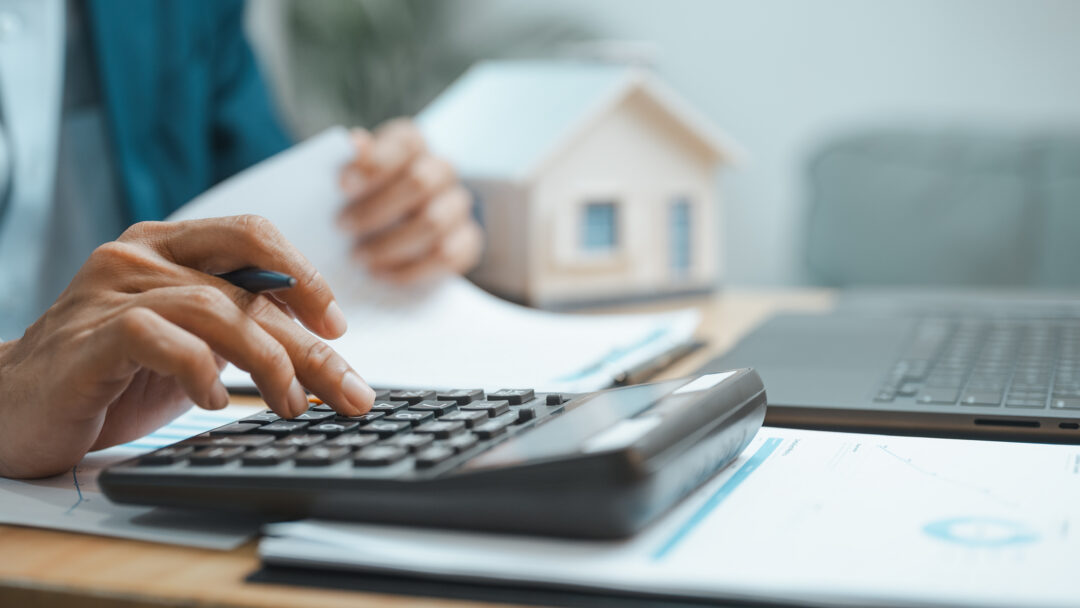 Person using a calculator while reviewing financial documents beside a laptop and model home.