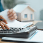 Person using a calculator while reviewing financial documents beside a laptop and model home.