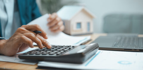 Person using a calculator while reviewing financial documents beside a laptop and model home.
