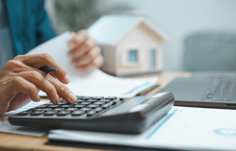 Person using a calculator while reviewing financial documents beside a laptop and model home.