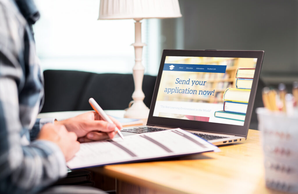 A person fills out a paper application beside a laptop displaying a college admissions website.