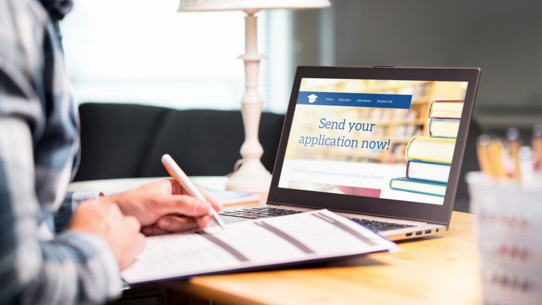 A person fills out a paper application beside a laptop displaying a college admissions website.