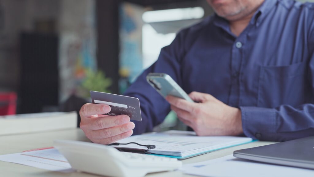 A man holds a credit card while using his smartphone at a desk covered with financial documents.