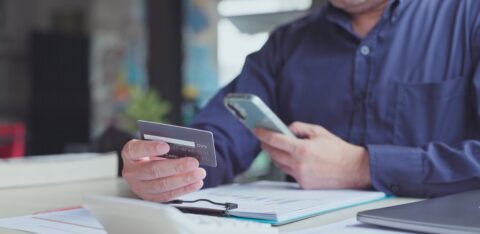 A man holds a credit card while using his smartphone at a desk covered with financial documents.