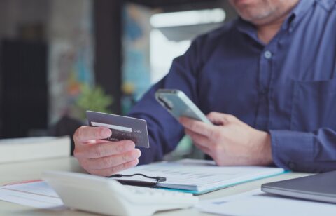 A man holds a credit card while using his smartphone at a desk covered with financial documents.