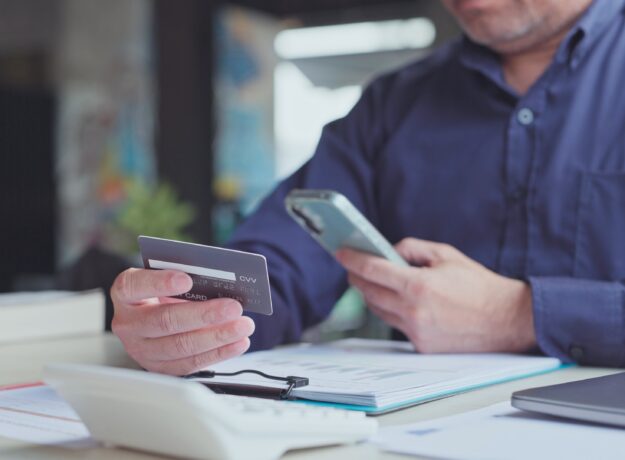 A man holds a credit card while using his smartphone at a desk covered with financial documents.