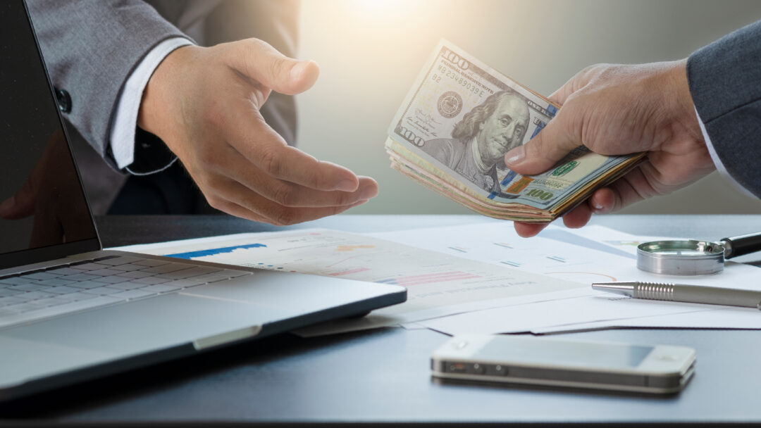 Close-up of one person handing a stack of hundred-dollar bills to another across a desk.