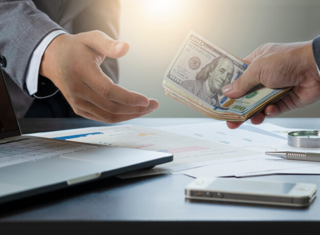 Close-up of one person handing a stack of hundred-dollar bills to another across a desk.