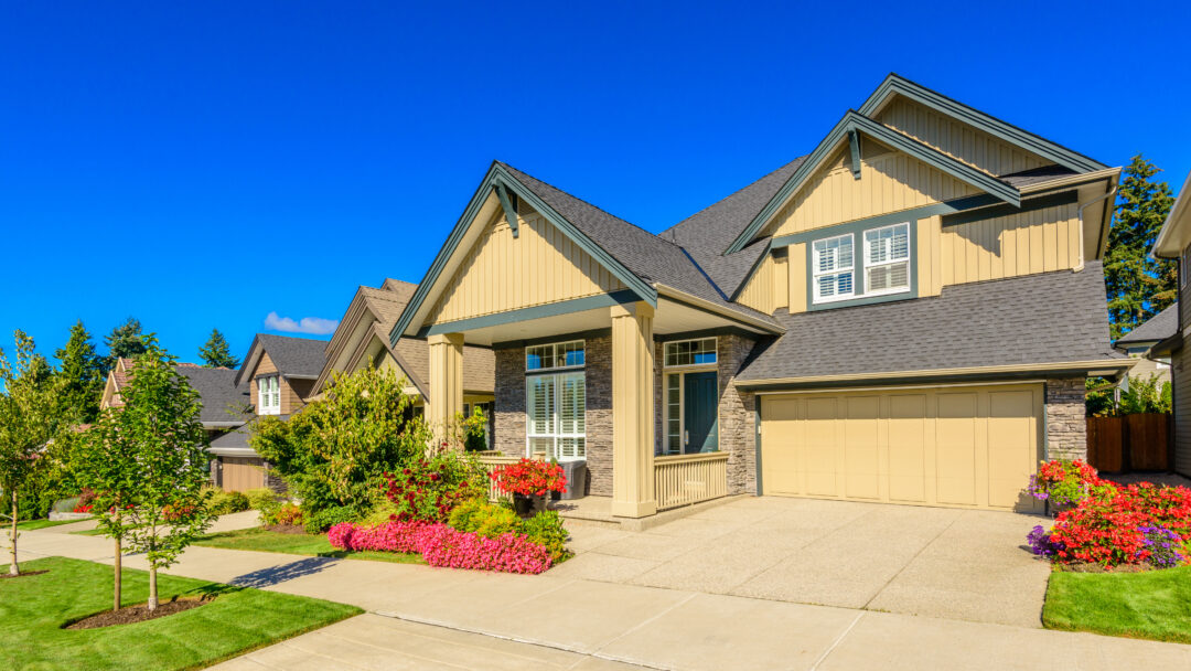 Large two-story suburban home with a two-car garage, landscaped front yard, and colorful flower beds.