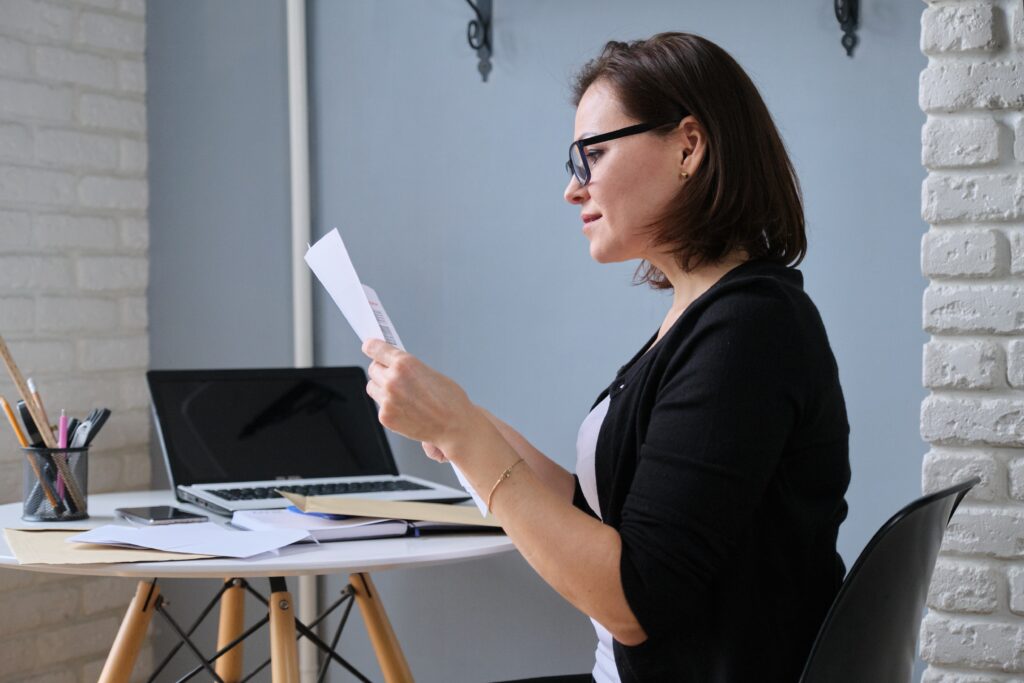 Woman wearing glasses reviewing paperwork while sitting at a desk with a laptop.