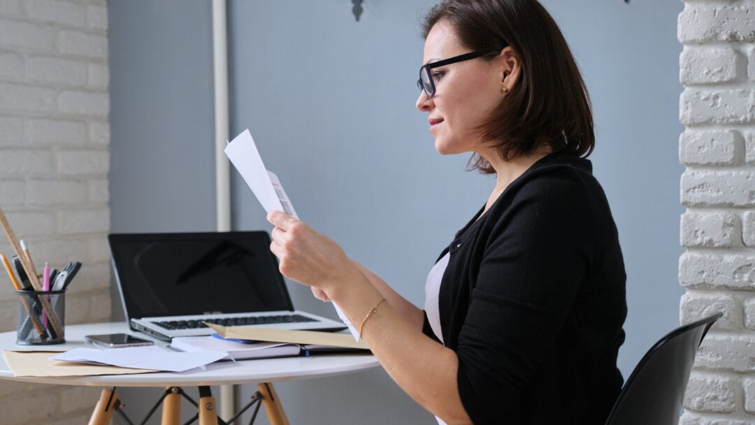 Woman wearing glasses reviewing paperwork while sitting at a desk with a laptop.