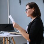Woman wearing glasses reviewing paperwork while sitting at a desk with a laptop.